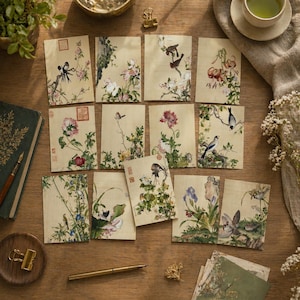 May include: A collection of rectangular paper cards featuring traditional East Asian style ink and watercolour illustrations of various birds, flowers, and foliage on a light beige background, arranged in a grid pattern on a wooden surface.