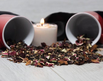 May include: A white candle with a red and white teacup spilling loose leaf tea onto a white wooden surface. The tea blend includes dried flowers, fruit, and herbs.
