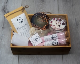 Könnte beinhalten: Eine Holzkiste gefüllt mit einem Selbstpflege-Geschenkset. Das Set enthält eine Tüte Tee mit der Aufschrift "Blushing Tea", einen kleinen weißen Beutel, eine Kerze mit Rosenblüten, zwei Glasröhrchen mit Badesalz und eine dekorative Achatplatte mit einem Kupferdraht-Baumdesign.