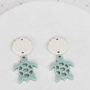 May include: Two earrings featuring a seashell and sea turtle design. The top portion is a cream-colored seashell shape, and the bottom is a light teal sea turtle with a honeycomb pattern on its shell. The earrings are displayed on a white surface.