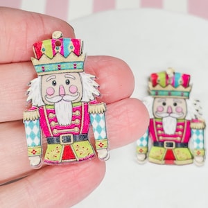 May include: Two flat nutcracker charms featuring a vibrant design with a pink jacket, teal and white diamond-patterned sleeves, a green base, and a crown. Each charm has a small hole at the top for hanging, showcasing a detailed illustration.