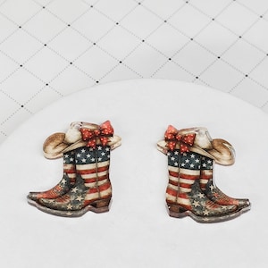 May include: Two decorative cowboy boot ornaments. Each boot features the American flag design with red and white stripes, blue stars, and a brown cowboy hat with a red bow. The boots are set against a white background.