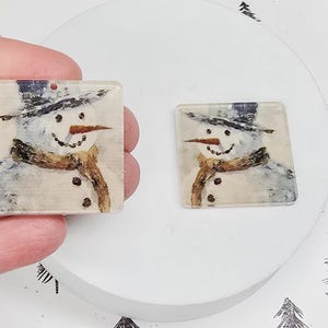 May include: Two square-shaped pendants featuring a watercolor snowman design. The snowman wears a black hat, a brown scarf, and has a carrot nose. The pendants are clear with a hole at the top for stringing.