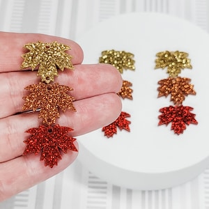 May include: Three sets of glitter maple leaf embellishments in gold, orange, and red. The leaves are arranged in a vertical line on a white surface.