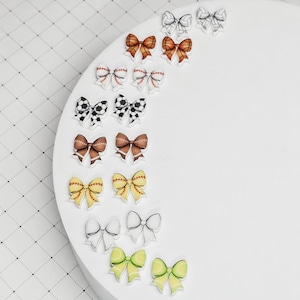 May include: A set of 12 pairs of colorful bow earrings with various sports-themed designs, including baseball, soccer, basketball, and softball.