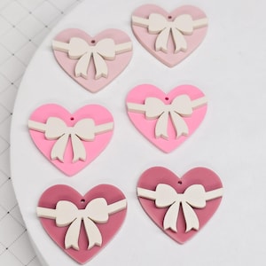 May include: Six heart-shaped ornaments in shades of pink and red. Each heart features a white bow and a horizontal white band. The ornaments are arranged on a white surface, perfect for Valentine's Day or other romantic occasions.