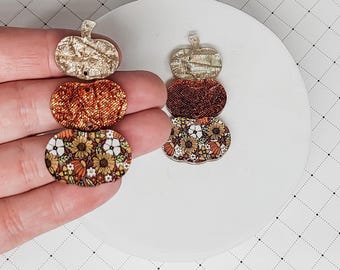 Op de afbeelding: Een set van drie gelaagde pompoenvormige oorbellen. De bovenste laag is goud, de middelste laag is oranje glitter en de onderste laag is een bloemenprint met witte, oranje en bruine kleuren.