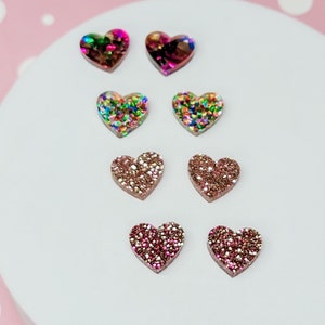 May include: Six pairs of heart-shaped earrings with glitter. The earrings are in shades of pink, red, and iridescent.
