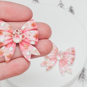 May include: Two pink and white bow-shaped resin ornaments with a hot chocolate and marshmallow design.