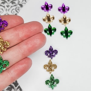 May include: Assortment of glitter fleur-de-lis charms in gold, green, and purple. The charms are displayed on a white surface.