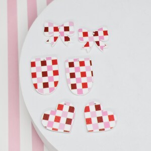 May include: A collection of decorative earrings in various shapes, including bows, ovals, and hearts. Each earring features a red, pink, and white checkered pattern. The earrings are displayed on a white surface with a pink and white striped background.