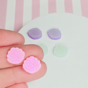 May include: Two pink glitter seashell shaped resin cabochons. The cabochons are on a hand, and there are other cabochons in the background in purple and light blue.