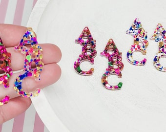May include: Four clear acrylic charms with a rainbow glitter embedded inside. The charms are shaped like the letters A, B, and C. Two additional charms are shaped like a seahorse.