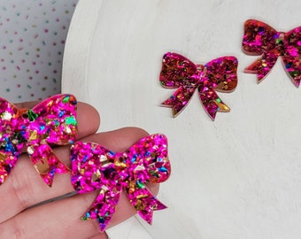 May include: Three pink glitter resin bow shapes with gold and iridescent glitter accents. The bows are on a white surface.