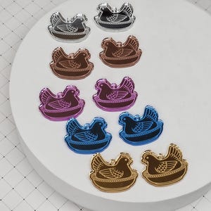 May include: A collection of colorful acrylic hen-shaped charms. The charms are in various colors, including silver, bronze, purple, blue, and gold. Each charm features a detailed hen design sitting in a nest. The charms are arranged on a white circular surface.