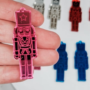 May include: A pink mirrored nutcracker ornament with a star on its hat. The ornament is held in a hand.