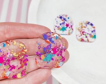 May include: A pair of clear resin earrings with a colorful splatter paint design. The earrings are oval shaped and have a translucent appearance.