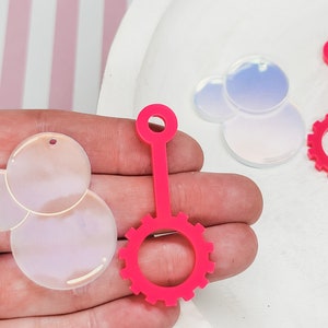 May include: Three pink plastic bubble wands. Two of the wands have a round bubble blower and a handle. The third wand has a round bubble blower with a gear-shaped handle.