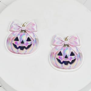 May include: Two iridescent pumpkin-shaped ornaments with a smiling jack-o'-lantern face and a pink bow on top. The pumpkins have a white border and are set against a white background, perfect for Halloween.