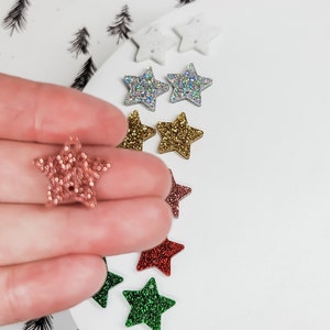 May include: Assortment of glitter star shapes in various colors, including gold, silver, red, green, and pink. The stars are arranged on a white surface.