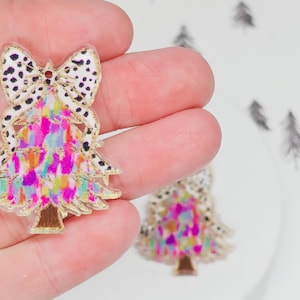 May include: A colorful Christmas tree-shaped charm with a bow on top. The bow has a white background with black spots. The tree is decorated with pink, yellow, and blue colors. The charm has a gold outline.
