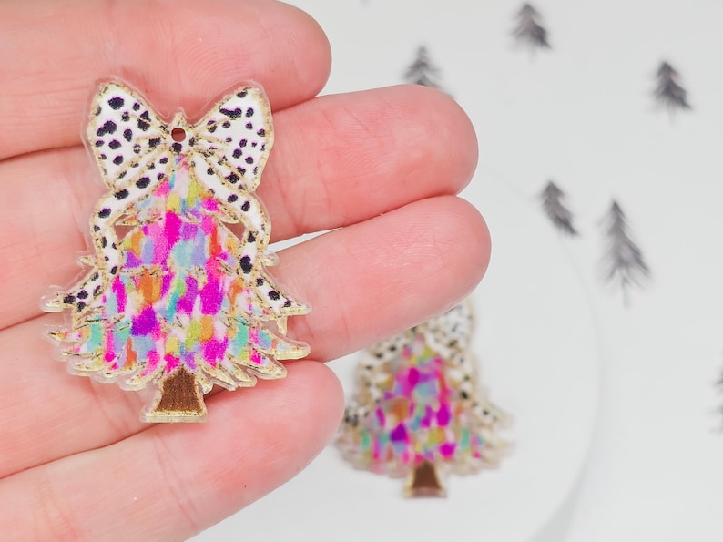 May include: A colorful Christmas tree-shaped charm with a bow on top. The bow has a white background with black spots. The tree is decorated with pink, yellow, and blue colors. The charm has a gold outline.