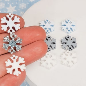May include: Assortment of snowflake charms in white, light blue, and silver. The charms are made of wood and have a glitter finish.