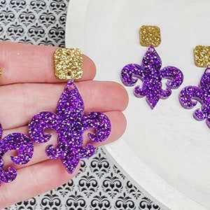 May include: A pair of purple glitter fleur-de-lis earrings with gold square accents. The earrings are dangling and have a hole at the top for a hook.