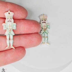 May include: Two small, hand-painted wooden nutcracker ornaments. The nutcrackers are in a traditional soldier design, with a light green uniform, a crown, and a mustache. One is held in a hand, the other rests on a white surface. Christmas decor.
