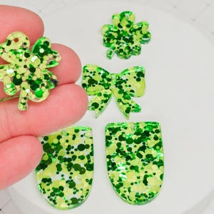 May include: A collection of green and yellow glitter resin charms in various shapes, including a four-leaf clover, a bow, and two shield-shaped pieces. The charms are on a white surface.