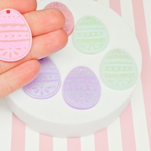 May include: Six pastel-colored Easter egg-shaped charms with a glitter finish and a white design. The charms have a hole at the top for hanging.