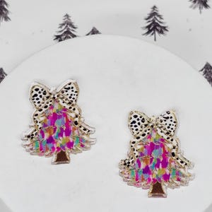 May include: Two Christmas tree-shaped earrings. Each earring features a bow with a black and white spotted pattern. The tree portion has a colorful, abstract design with pink, purple, and blue hues. The base of the tree is brown.