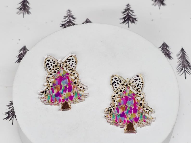 May include: Two Christmas tree-shaped earrings. Each earring features a bow with a black and white spotted pattern. The tree portion has a colorful, abstract design with pink, purple, and blue hues. The base of the tree is brown.