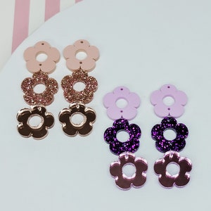 May include: Two pairs of flower-shaped earrings. One pair features a light pink top, glittery rose gold center, and rose gold bottom. The other pair has a light purple top, glittery purple center, and rose gold bottom. The earrings are on a white surface.
