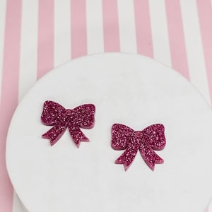 May include: Two glittery magenta bow-shaped embellishments. The bows are on a white circular surface against a pink and white striped background. The bows have a sparkly texture and are symmetrical.