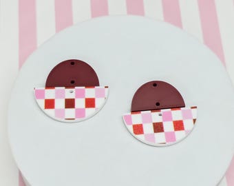 May include: A pair of earrings with a burgundy half-circle top and a checkered pattern in red, pink, and white. The earrings are on a white surface with a pink and white striped background.