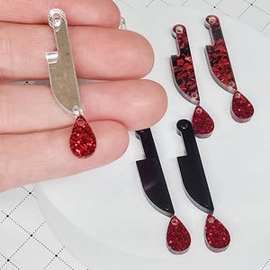 May include: A collection of novelty earrings featuring a knife design with a red glitter teardrop. Some knives are silver, others are black with red glitter accents. The earrings are designed for pierced ears.