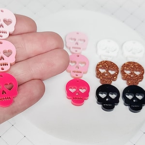 May include: Assortment of colorful skull charms with heart-shaped eye holes. The charms are made of acrylic and come in pink, white, black, and copper.