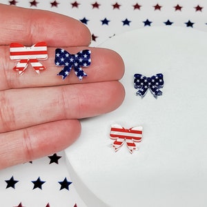 May include: Small, decorative bow-shaped embellishments in red, white, and blue. Two bows feature red and white stripes, while the others display a blue background with white stars. These patriotic-themed bows are ideal for crafting or adding a festive touch.