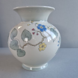 May include: A white ceramic vase with a round body and a flared rim. The vase is decorated with a floral pattern featuring blue, yellow, and red flowers, along with green leaves and vines. The base and rim have a light blue trim.
