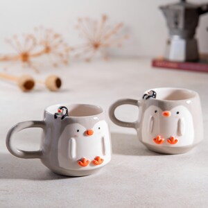 May include: Two gray ceramic mugs with penguin designs. Each mug has a small penguin perched on the rim. The penguins have orange beaks and feet.