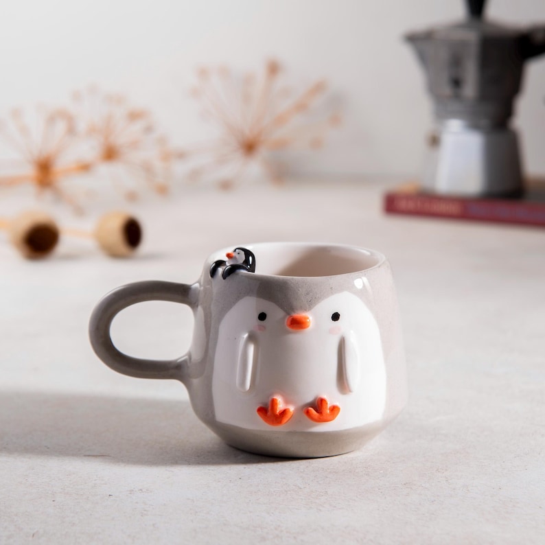 May include: A gray ceramic mug shaped like a penguin with a smaller penguin on its back. The mug has orange feet and a white face with black eyes.
