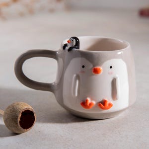 Puede incluir: Una taza de cerámica gris con forma de pingüino con un pingüino más pequeño posado en el borde. La taza tiene detalles naranjas en los pies y el pico del pingüino.