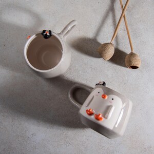 May include: Two ceramic mugs with penguin designs. One mug is gray with a penguin on the side. The other mug is white with two penguins on the side.