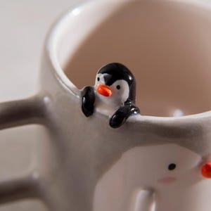 May include: A ceramic mug with a penguin design. The penguin is peeking over the edge of the mug, with its head and wings visible. The mug is gray and white.