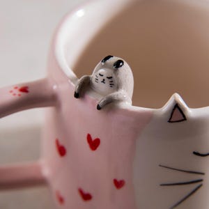 May include: A pink and white ceramic mug with a cat design. The mug has a cat's head on one side and a cat's tail on the other. The cat's head is 3D and has a pink nose and black eyes. The mug is decorated with red hearts.