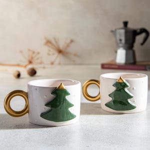 May include: Two white ceramic coffee mugs with gold handles and a green Christmas tree design. The mugs are on a white surface.