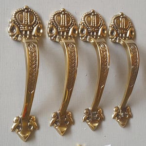 May include: Four brass-colored cabinet handles with a crown design at the top and a curved handle with leaf detailing. A small pile of gold screws is below the handles. The handles are on a light gray surface.