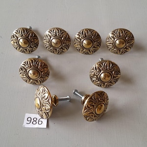 May include: Ten gold-toned cabinet knobs with a decorative sunburst design. The knobs have a round shape and a raised center. The knobs are arranged in a circle on a white surface.