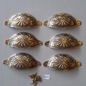 May include: Six antique brass-toned drawer pulls with an ornate, symmetrical design. Each pull has a curved, half-moon shape with decorative detailing. Small screws are included. The pulls are arranged on a light gray surface.